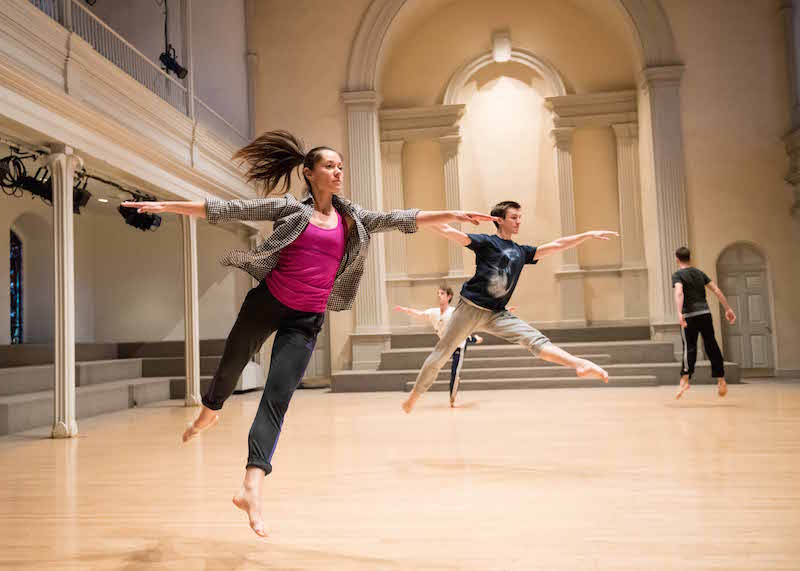 Melissa Toogood in mid arabesque jump while other dancers mirror her in the background. They all wear practice clothes